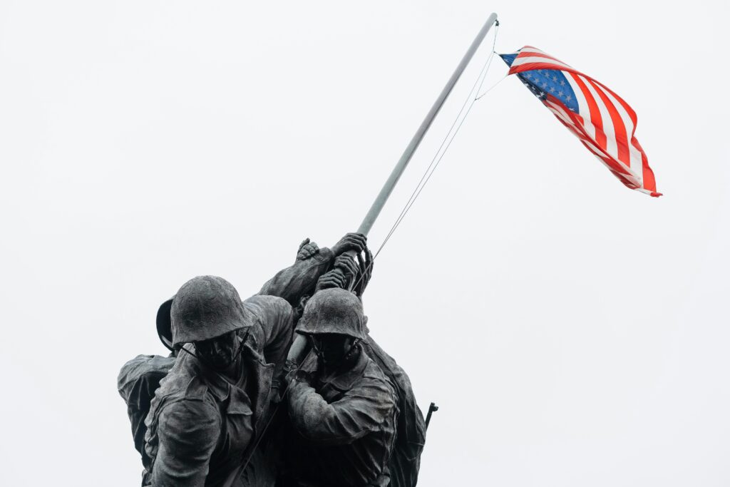 A close-up of a statue depicting soldiers raising an American flag captures the essence of unity and perseverance. The flag waves triumphantly above the determined figures against a clear, pale sky, reminiscent of scenes often shared by Firearm Blog that honor valor and resilience.