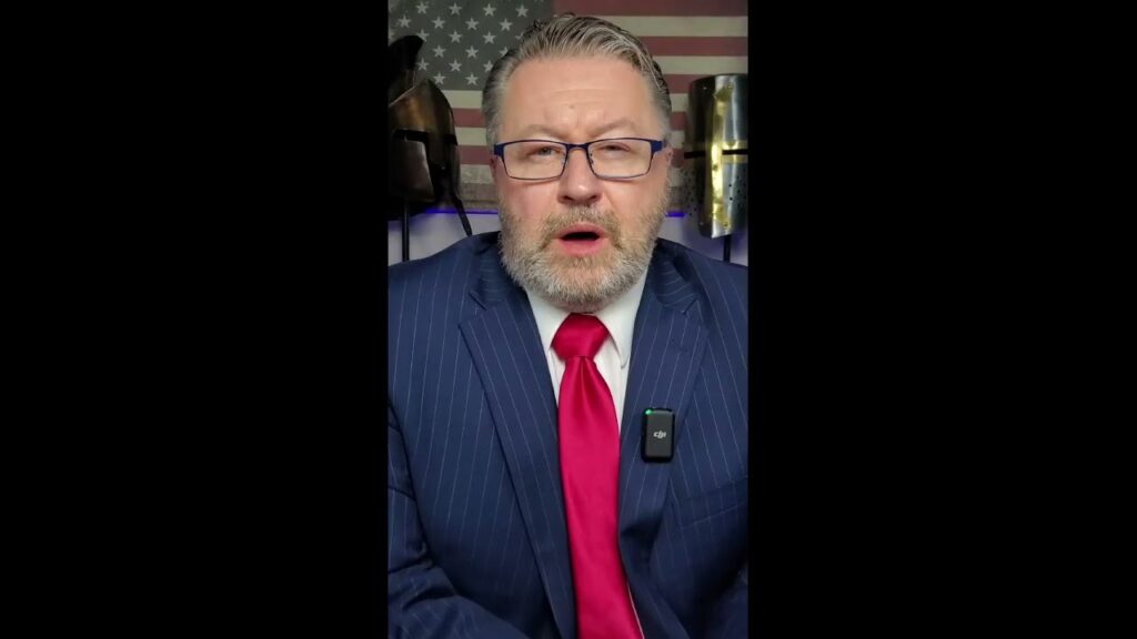 Man with gray hair and beard wearing a blue pinstripe suit, red tie, and glasses speaking in front of a U.S. flag. He has a microphone clipped to his lapel.