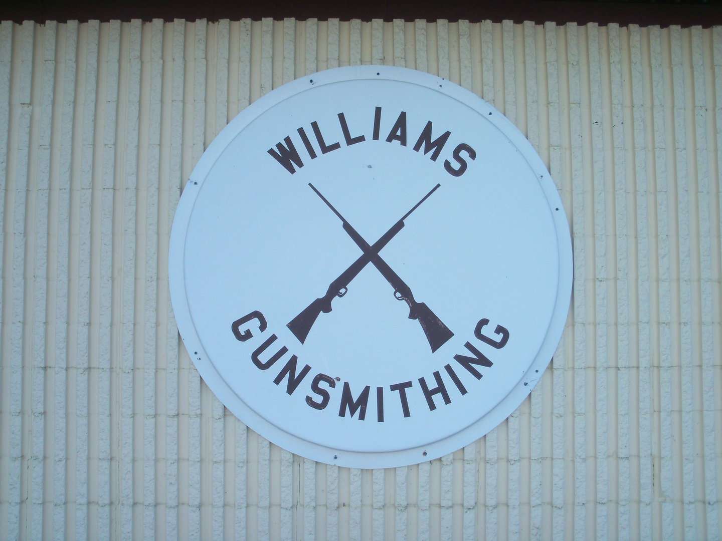 A white circular sign on a ribbed wall features two crossed rifles in the center. The text "Williams Gunsmithing" arches above and below the rifles in bold, black letters.