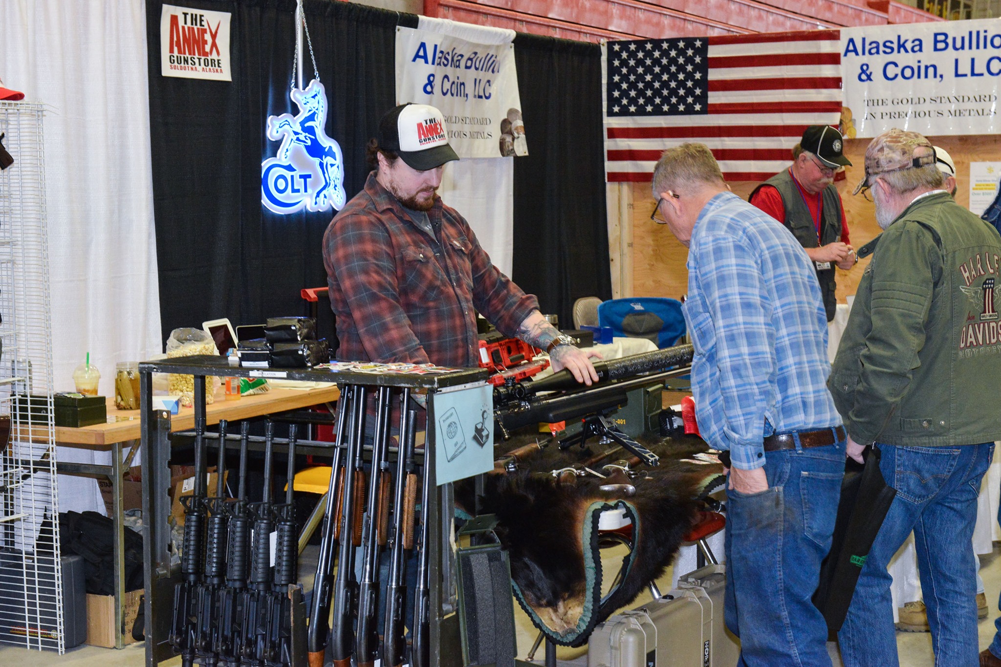 People engage at a gun show booth with firearms displayed. A man in a plaid shirt behind the counter talks to two men examining a gun. American flags and promotional signs are visible in the background.