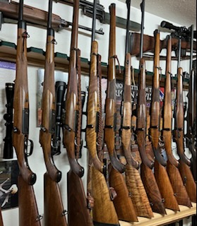 A row of hunting rifles with wooden stocks are lined up vertically against a wall, with scopes mounted on some. There are various accessories and equipment visible in the background.