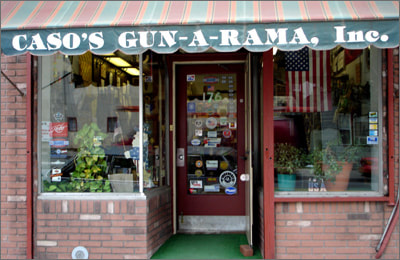 Storefront of "CASO'S GUN-A-RAMA, Inc." with a striped green awning, brick exterior, glass door covered in stickers, and potted plants inside. An American flag hangs in the window.