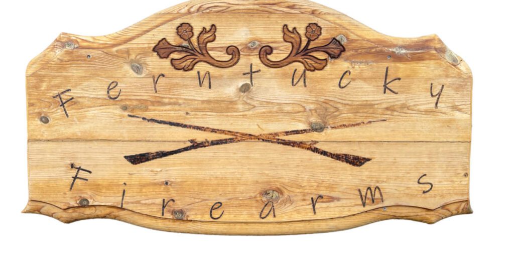 A wooden sign with engraved floral designs at the top and the words "Ferntucky Firearms" in a handwritten style, featuring two crossed rifles in the center.