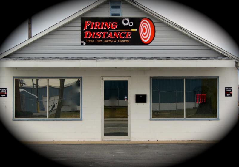 A small, single-story gun shop with a sign reading "Firing Distance" and a target logo above the entrance. The building is white with large windows on either side of a glass door.