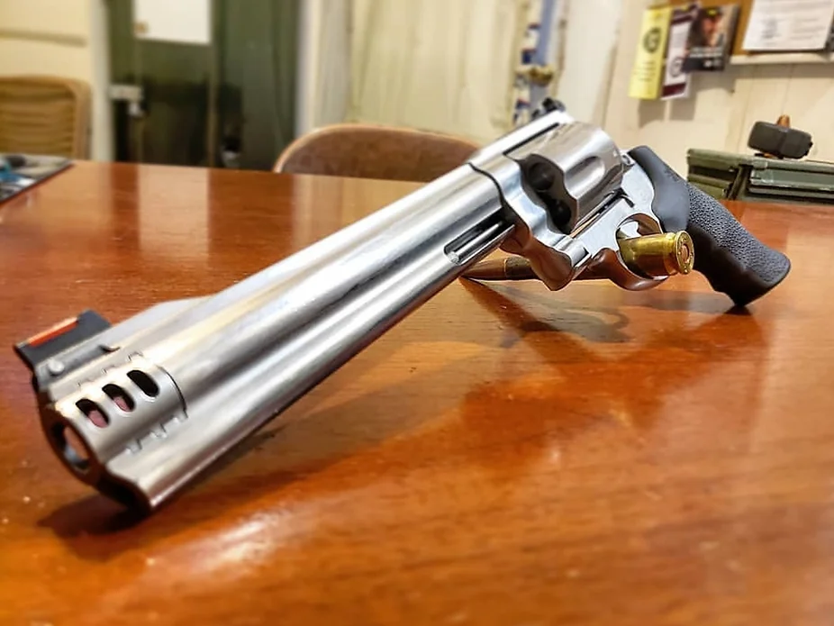 A large silver revolver with a long barrel and a black grip rests on a polished wooden table, with a bullet inserted in its cylinder. Background shows an office setting with chairs and papers.