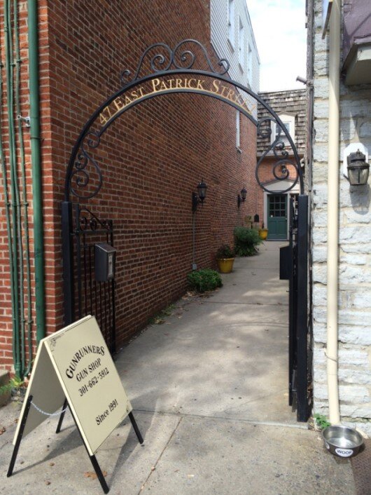 A wrought iron archway leads to a narrow alley lined with brick walls. A sign beside the alley reads "Gunrunners Gun Shop" with a phone number, and there are yellow potted plants along the path.