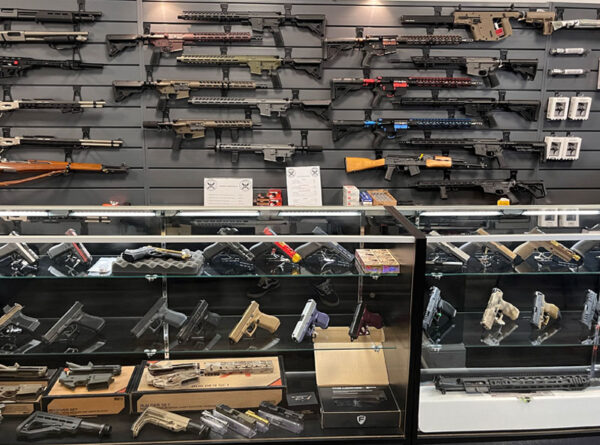 A gun store display with various rifles mounted on a wall and multiple handguns arranged in glass cases below, along with firearm accessories and ammo boxes.