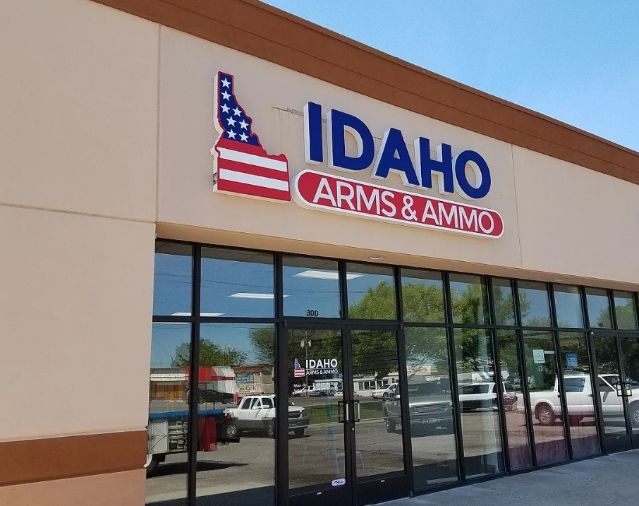 Storefront of "Idaho Arms & Ammo" with a sign featuring a red, white, and blue design resembling the state of Idaho. The reflection of cars and trees is visible in the large glass windows.