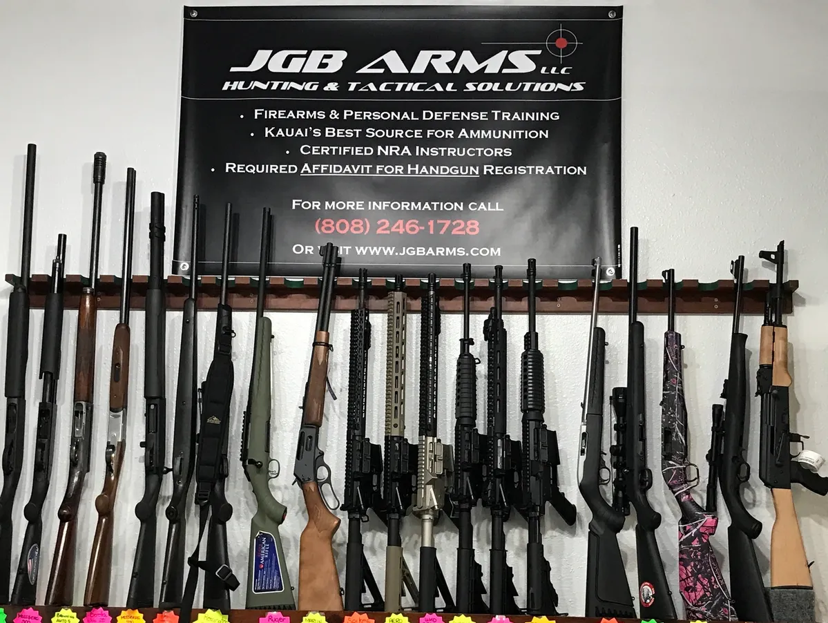 A display of various rifles and shotguns lined up against a wall beneath a banner. The banner reads: "JGB ARMS LLC Hunting & Tactical Solutions." Contact details and additional services are listed on the banner.