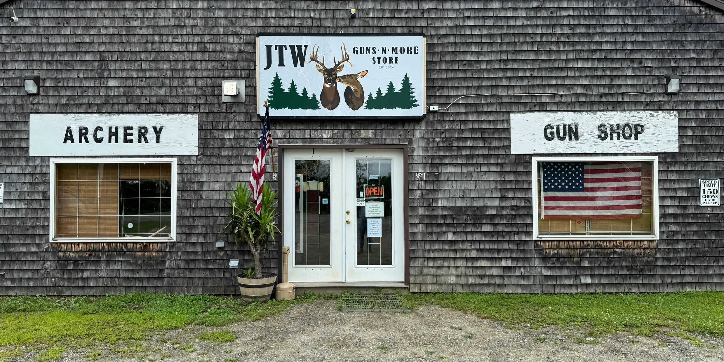A rustic wooden storefront with signs reading "Archery" and "Gun Shop," an American flag in the right window, potted plants by the entrance, and a sign above reading "JTW Guns N' More Store" with a moose logo.