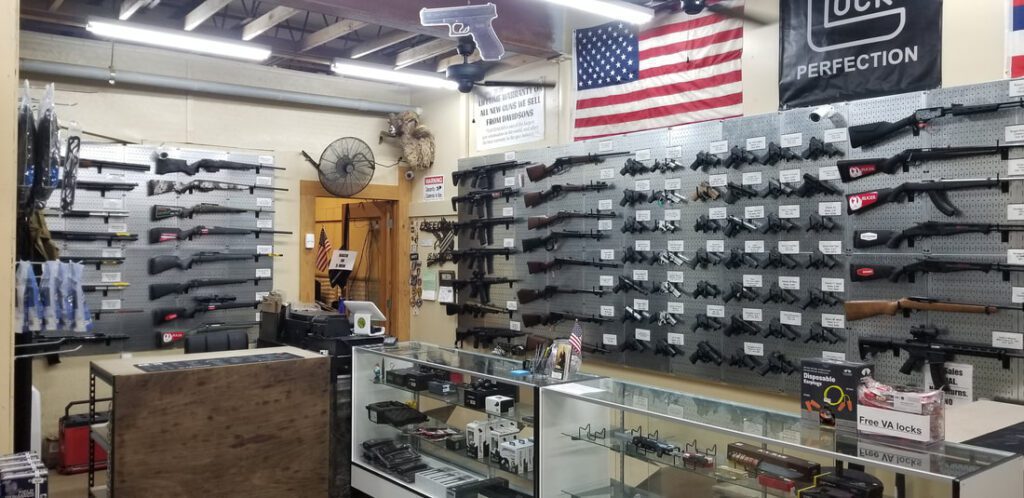 A gun store interior features multiple rifles and handguns displayed on pegboards. A glass counter showcases various accessories. An American flag and a large Glock poster hang on the wall. The store has a rustic wooden ceiling and cluttered decor.