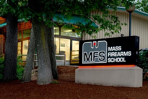 Exterior view of Mass Firearms School building with large illuminated MFS sign near entrance, surrounded by trees and landscaping.