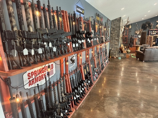 Single Category 5 Rows of rifles are displayed on wooden racks along the wall of a gun store, with signs for Springfield Armory and other brands visible. The store has polished concrete floors and mounted animal heads on the far wall.