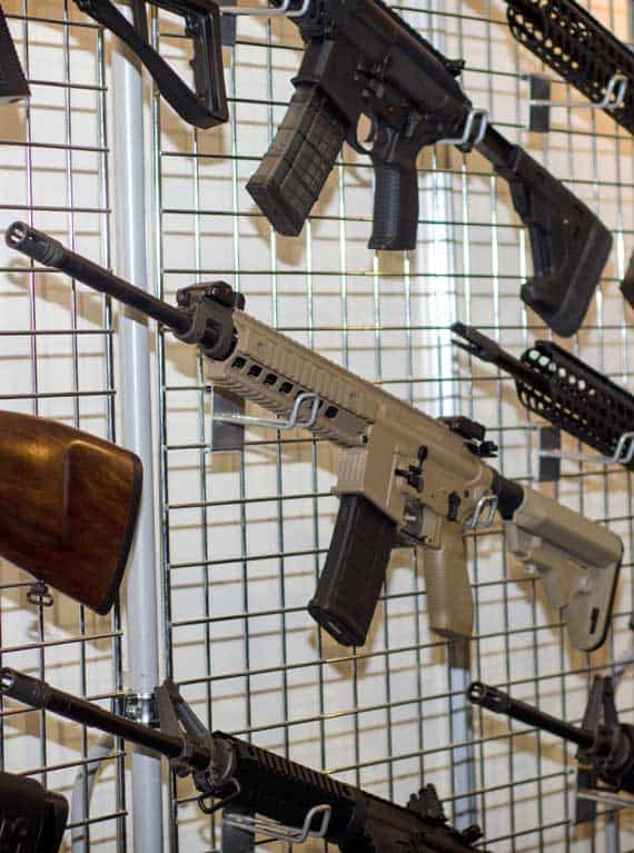 A display of rifles mounted on a wire grid wall. The central focus is a tan-colored rifle with a black magazine. Other black rifles are arranged nearby, showcasing various designs and features.