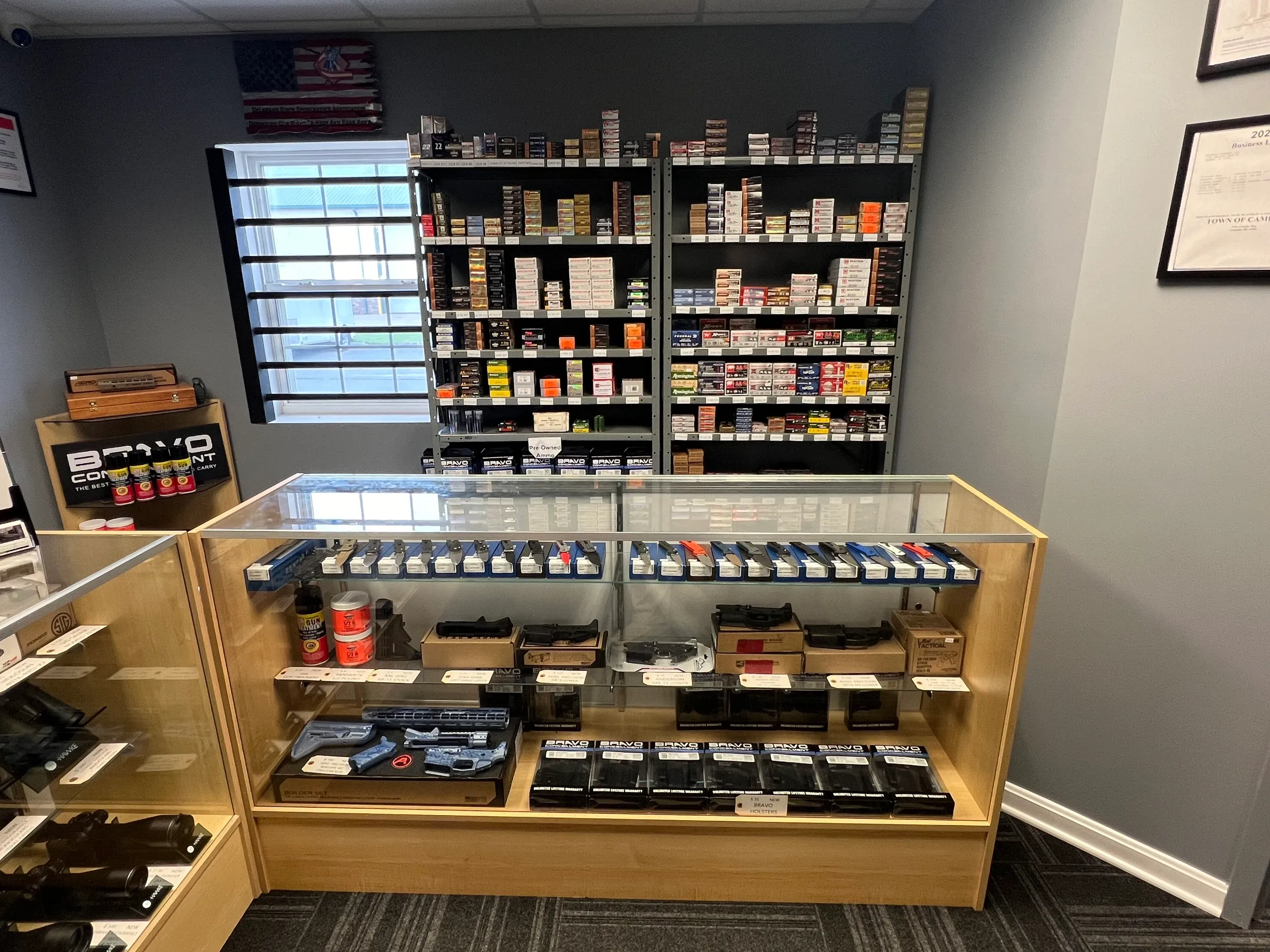 A store display featuring shelves filled with various ammunition boxes behind a glass counter. The counter displays more ammunition in organized rows and stacks, with additional firearm accessories beneath. Framed certificates hang on the nearby wall.