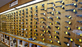 Missouri Firearms Training 6 A wooden wall display in a store shows dozens of handguns, each with a yellow price tag. Below the guns, shelves hold boxes and accessories. The display stretches across the length of the wall.