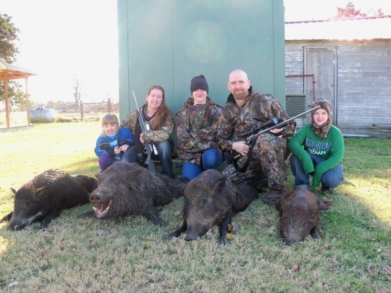 A group of people wearing camouflage clothing are posed kneeling on grass. They are holding rifles and are surrounded by several wild boars lying on the ground. A green structure and a wooden building are in the background.
