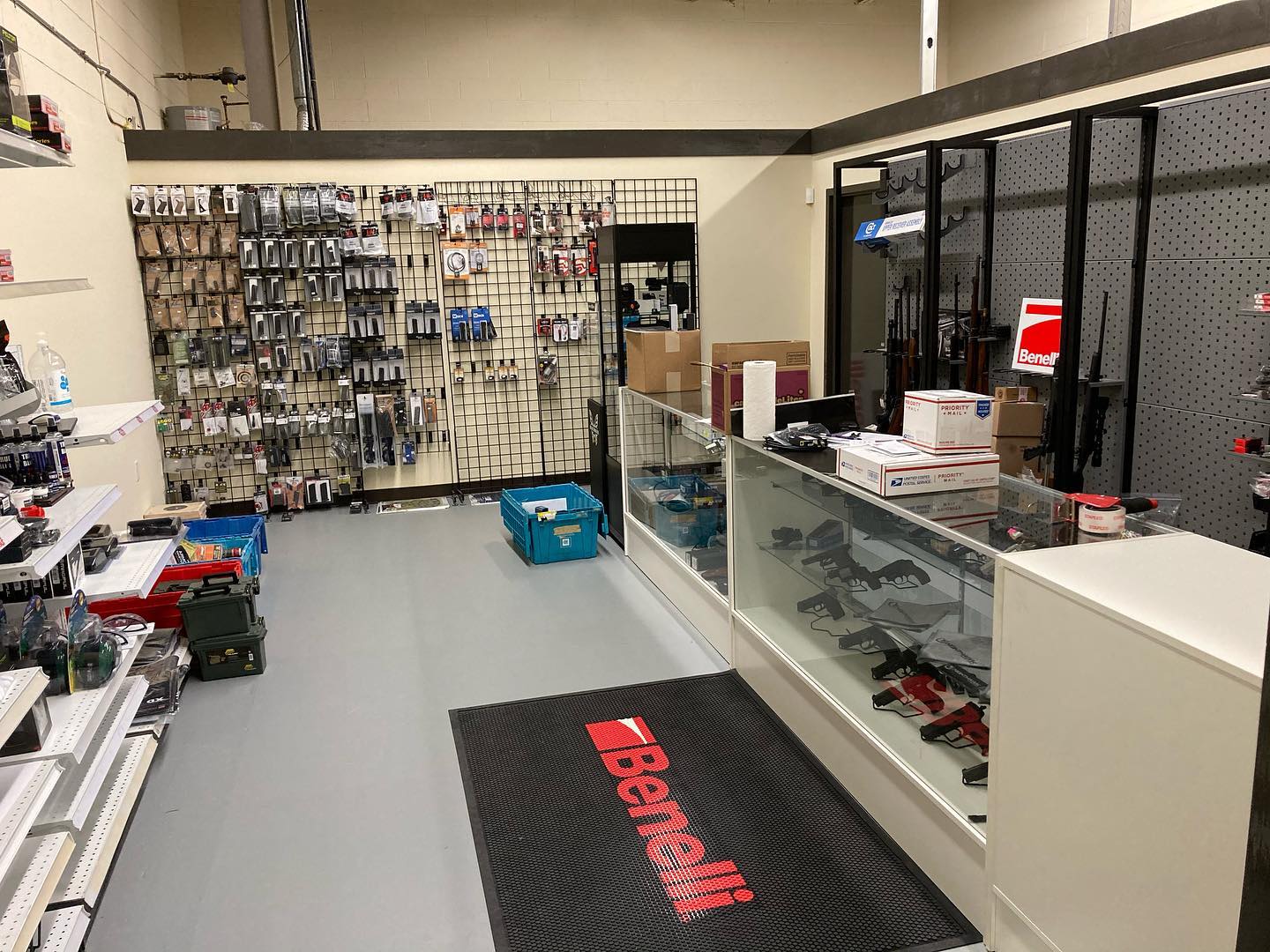 Single Category 1 A gun shop interior with firearms and accessories displayed on shelves and racks, glass counters with handguns and boxes, and a black floor mat with a red Benelli logo near the entrance.