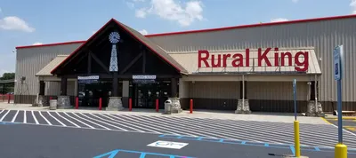 Storefront of a Rural King building with a beige exterior and red lettering. A small windmill decoration is above the entrance. The parking lot in front is marked with blue and white lines for handicapped parking spaces.