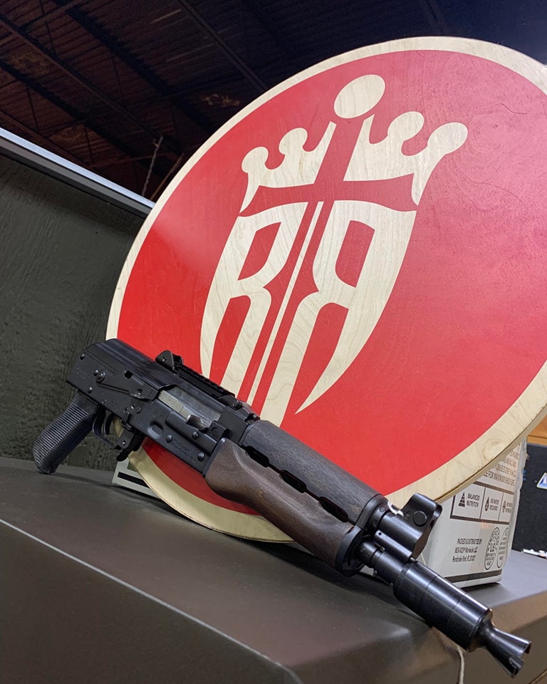 A short-barreled black rifle with a wooden foregrip is propped against a circular red and white sign featuring a stylized crown and shield design, with the letters "BR" in the center.
