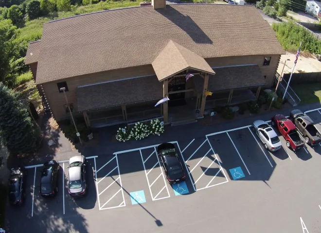 Aerial view of a brown building with a pitched roof, surrounded by trees. In front, a parking lot with several cars and marked accessible parking spaces. Flags are displayed near the entrance. Shadows cast on the pavement indicate a sunny day.