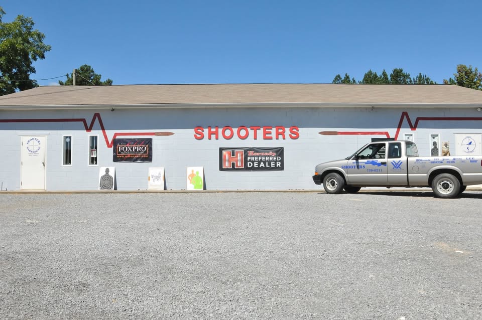 A one-story building with a sign that reads "Shooters" above the entrance. A pickup truck is parked in front. Various signs and targets are displayed along the structure's light gray exterior. Trees are visible in the background under a clear blue sky.