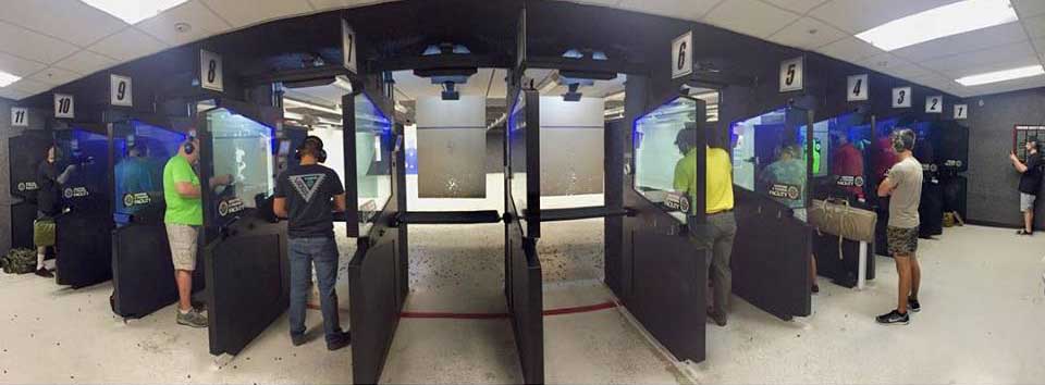 People in a shooting range wearing protective earmuffs, aiming guns at targets. The indoor range has individual stalls with partitions for each participant, and overhead lighting. Targets are visible in the distance, and spent casings litter the floor.