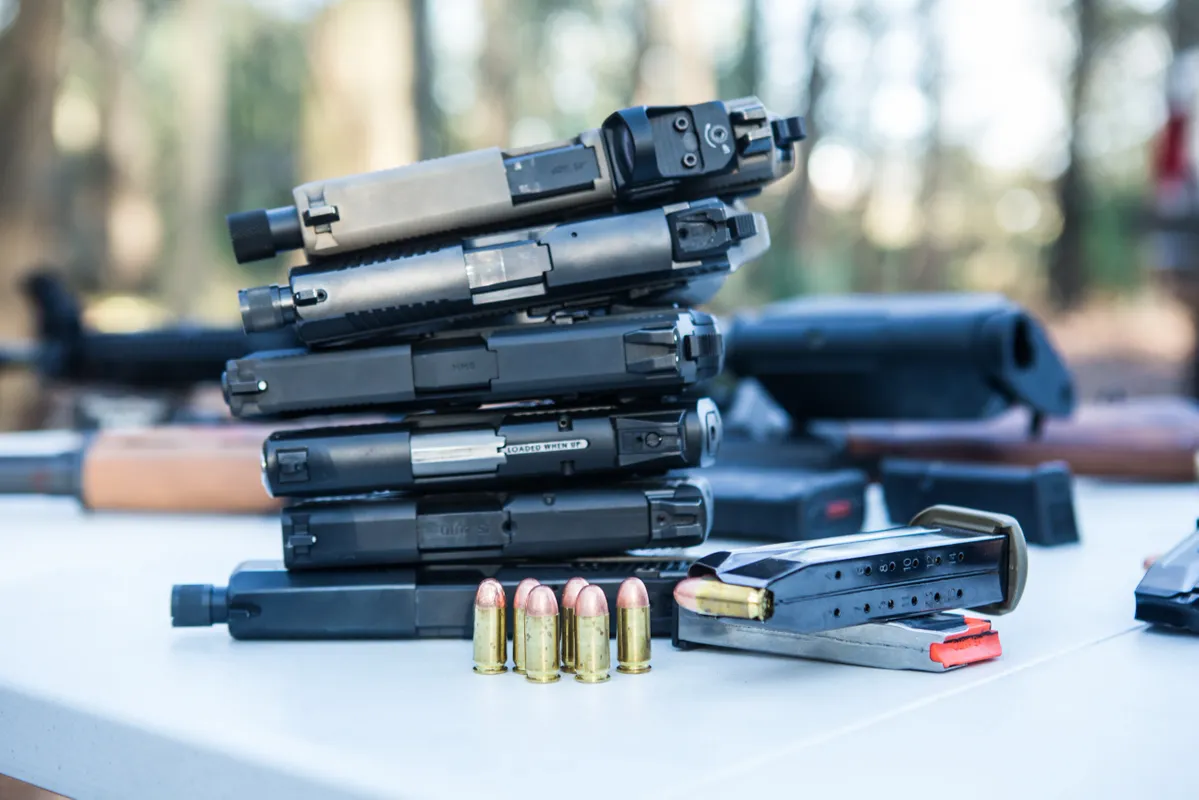 A stack of five handgun slides is arranged on a white table outdoors. In front of the stack are six bullets and a loaded magazine. The background is blurred, showing trees and additional firearms.