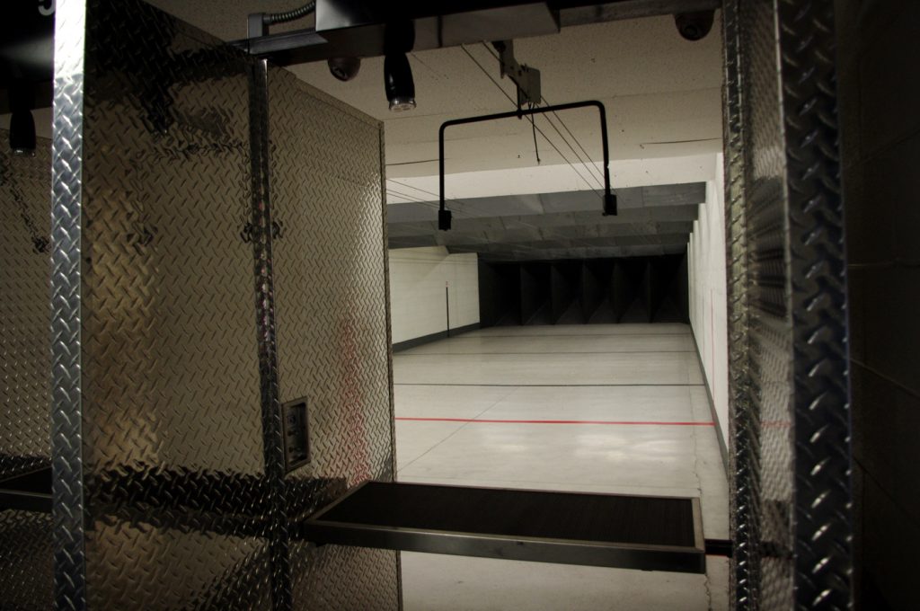 Indoor shooting range with empty shooting lanes, metal partitions, and a target track system visible, leading down a long, well-lit corridor.
