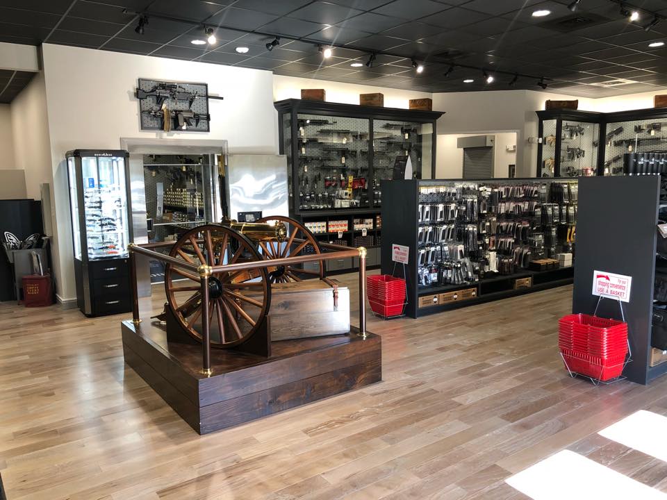 Single Category 6 A modern gun store interior with wooden floors, display cases of firearms and accessories, and a large antique cannon on a wooden platform in the center. Red shopping baskets are stacked near the entrance.
