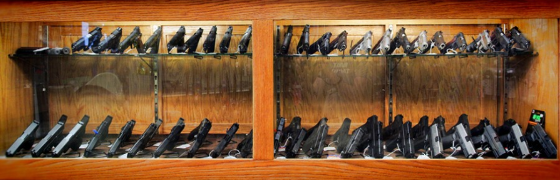 A glass display case with two sections, each filled with various handguns arranged in rows on two shelves, set against a wooden frame background.