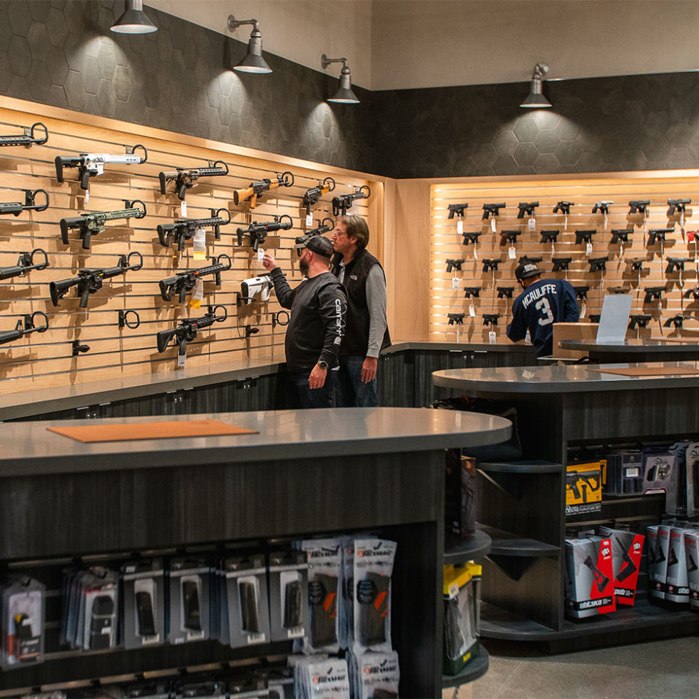 Two people inside a gun store examine rifles displayed on a wall. The store displays various firearms and accessories on shelves and racks. One person points at a rifle, and a jersey with "SCHULTE 3" is visible in the background.