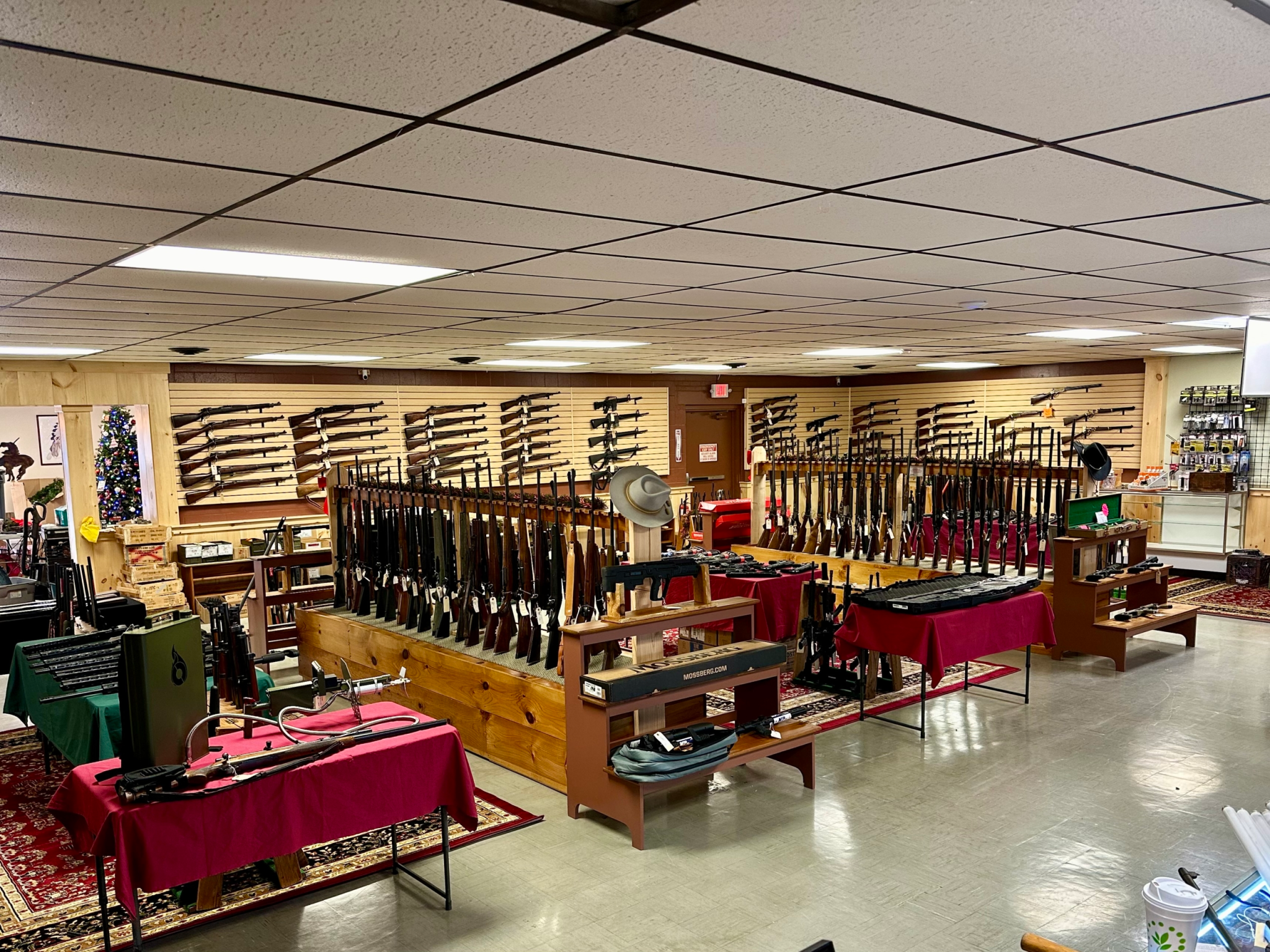 Single Category 2 A gun shop interior with rifles and shotguns displayed on wall racks and wooden stands, tables covered with red cloths, and various accessories and items on shelves around the room.