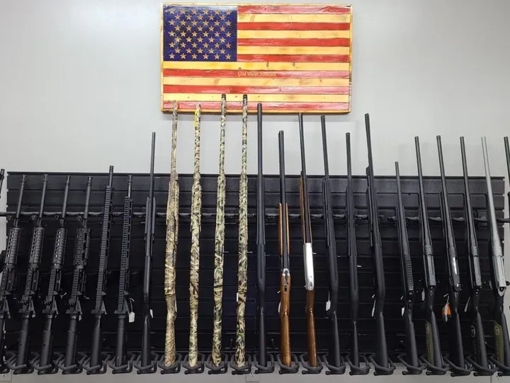 Single Category 3 A display of various rifles and shotguns is arranged upright on a rack beneath a wooden American flag on a white wall.
