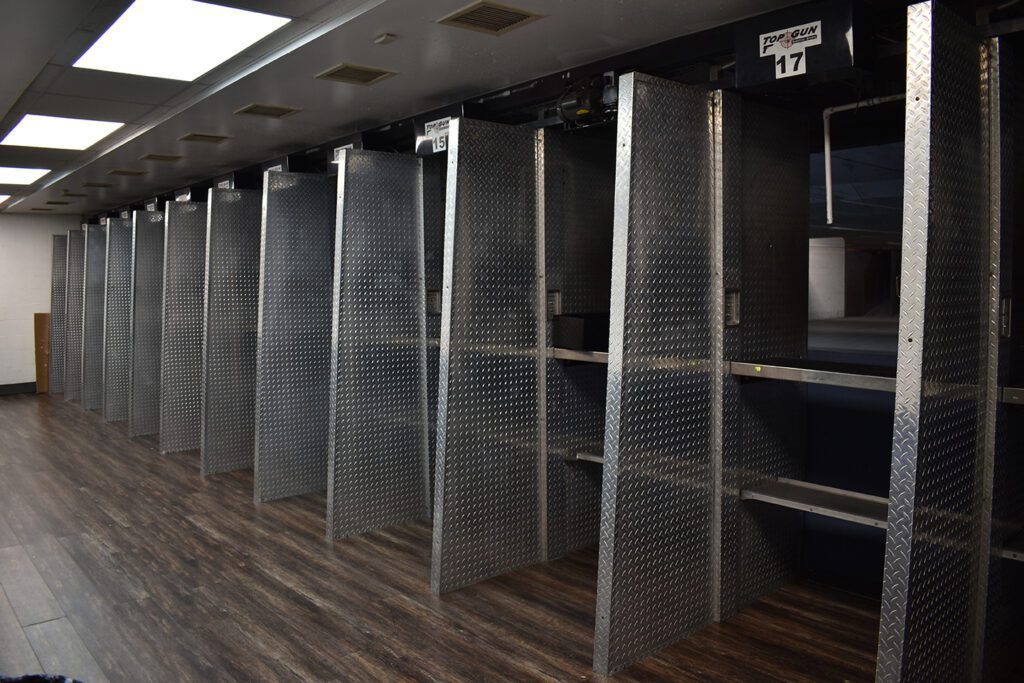 A row of empty shooting range stalls with metallic dividers, numbered signs above each booth, and dark wood flooring. The stalls face towards a back wall with targets not visible in the image.