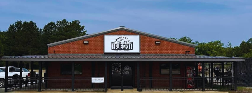 A single-story red brick building with a sign reading “Trading Co. True Grit” above the entrance, featuring a covered porch, black fencing, and trees in the background under a blue sky.