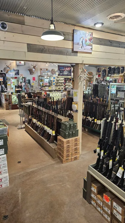 Single Category 5 Interior of a firearm store with rifles displayed on stands and shelves. Boxes of ammunition are stacked nearby. The store has wooden walls, a corrugated metal ceiling, and various hunting accessories on display. Hanging lights illuminate the space.