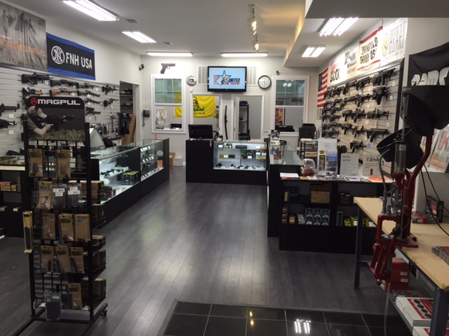 Interior of a gun store featuring display cases with firearms on the walls. The room is brightly lit with wooden flooring, and there are various accessories and products on shelves and racks, along with a counter and a television screen.