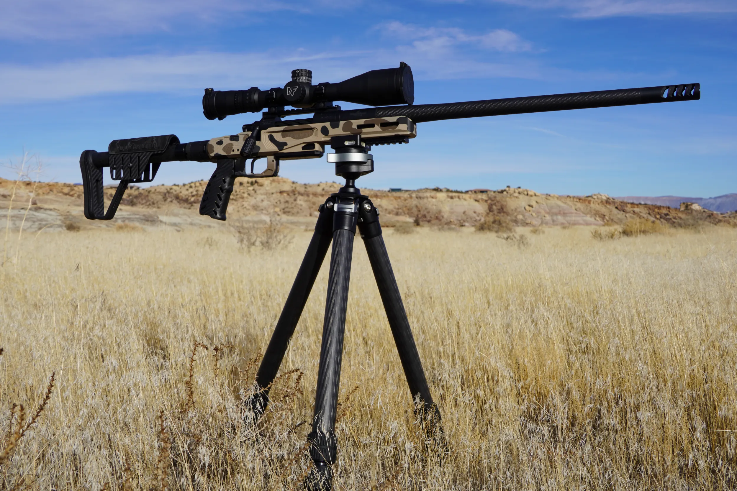A rifle with a camouflage pattern and scope is mounted on a tripod, set in a dry grass field under a blue sky with light clouds. Rocky terrain is visible in the background.