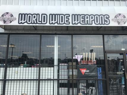 Storefront of "World Wide Weapons" with a sign displaying the name. The front features bars on windows and visible under the sign are some items inside the store, including a red Trijicon sign.
