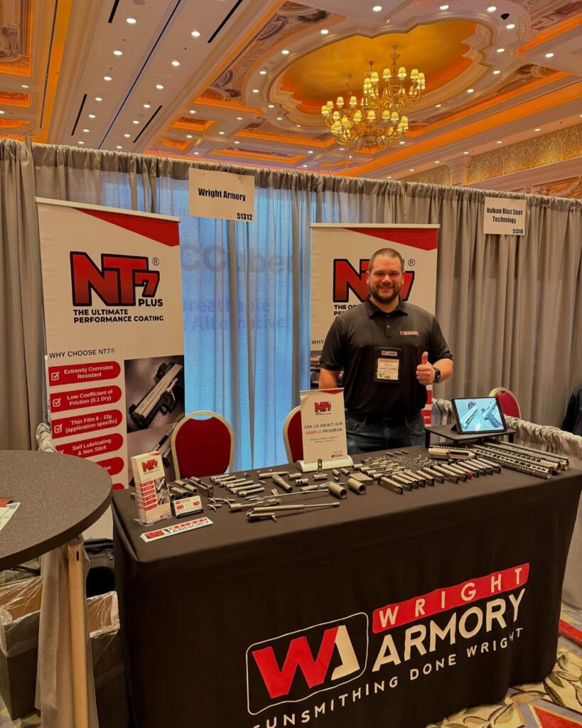 A man stands smiling at a trade show booth for Wright Armory. The booth features banners for NT7 Plus coating and displays various firearm components. The background shows an elegant, well-lit room with multiple booths and red chairs.
