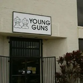 A building entrance with a sign above reading "Young Guns." The sign features two cartoon-style characters. The entrance has a black railing and plants are visible to the right of the doorway.