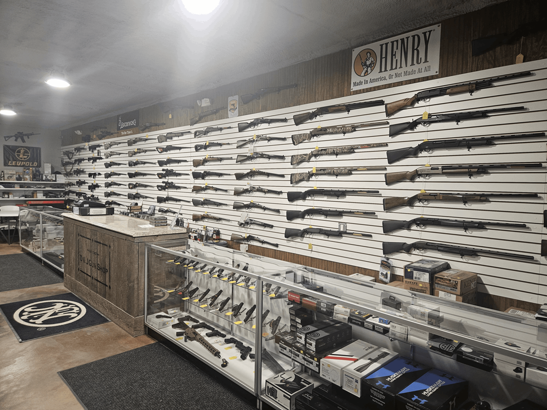 Single Category 2 A gun store interior with rifles displayed on wall racks, glass display cases filled with handguns and accessories, and signs for Henry and Leupold brands visible above and on the counter.
