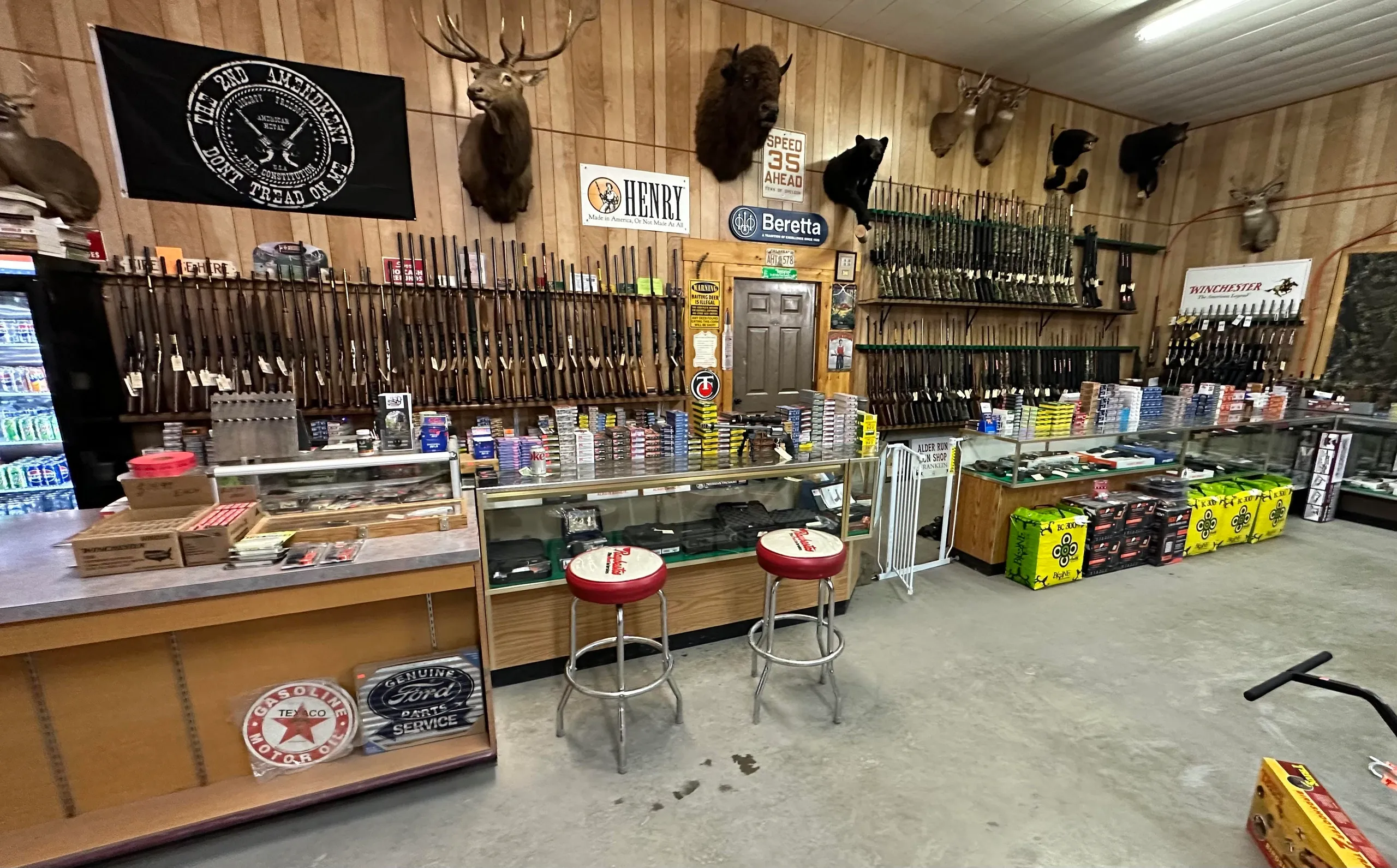 Single Category 3 A gun shop interior with rifles displayed on wooden walls, ammunition boxes on glass counters, mounted deer heads, black stools, vintage signs, and snacks and drinks along the side.
