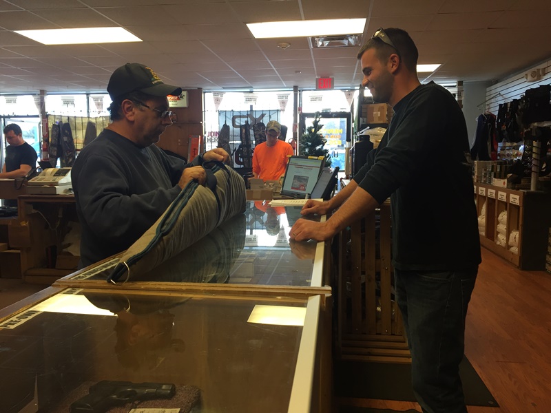 Two men stand at a store counter, one unrolling fabric while the other looks on. Shelves with various items and other customers are visible in the background. Natural light streams in from large front windows.