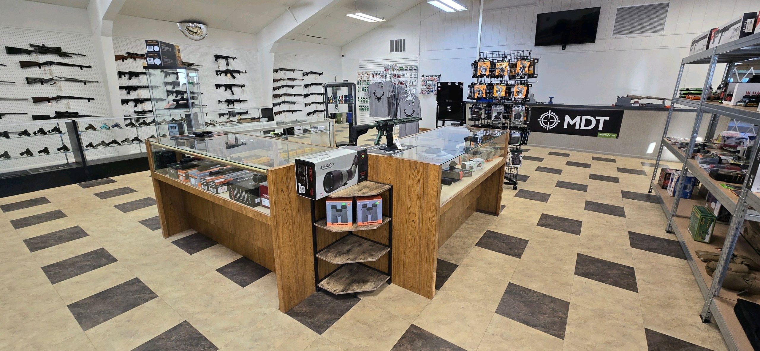 A gun shop interior with display cases of firearms and accessories, rifles mounted on the walls, shelves with gear, and a counter labeled "MDT" near the back, all on a checkered tile floor.