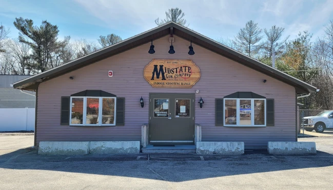 Firearms Training Directory 14 A single-story building with a brown exterior and a sign reading "Mustard Gun Company Indoor Shooting Range" above the entrance, situated in a parking lot with trees in the background.