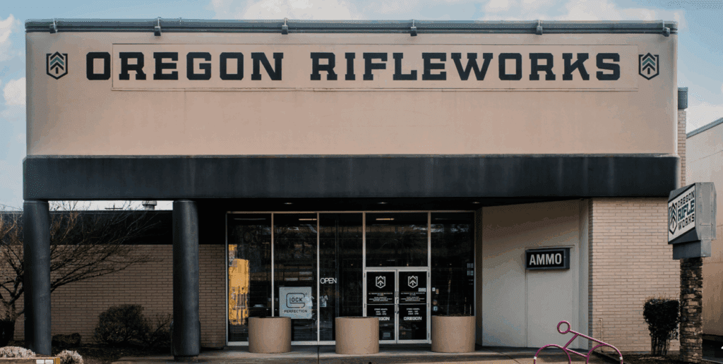 A storefront with a large sign reading "Oregon Rifleworks" above glass doors. The entrance has "AMMO" and "OPEN" signs, and a pink bicycle rack is visible near the entrance.