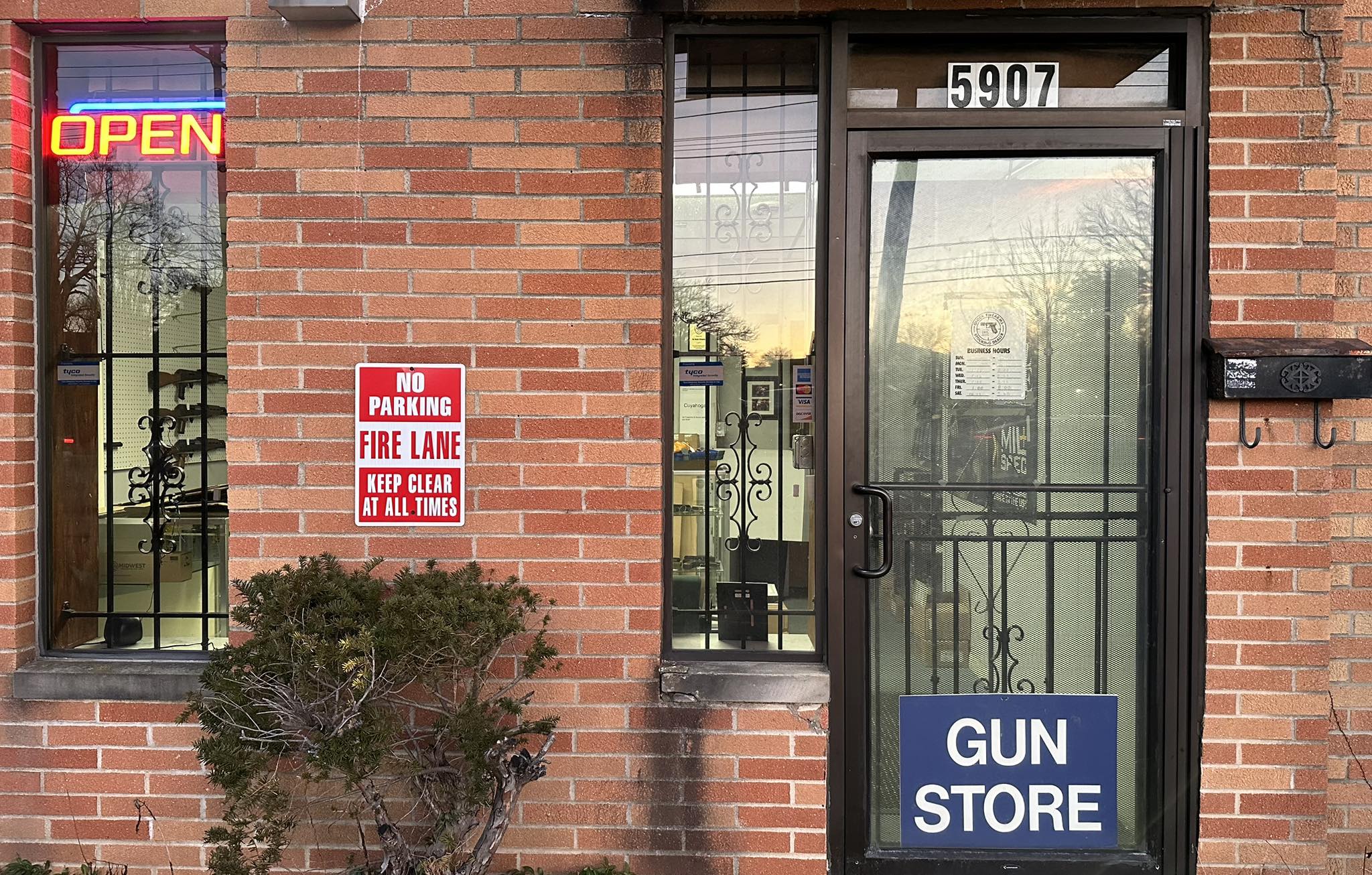 A brick building with a glass door displaying a "GUN STORE" sign and street number 5907. A neon "OPEN" sign and a red "No Parking Fire Lane" sign are visible; a small shrub is near the entrance.