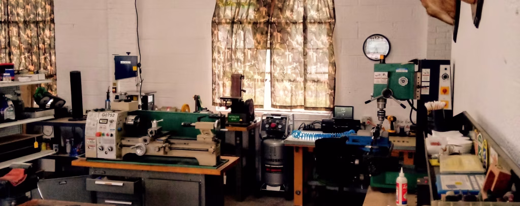 A workshop with various machines and tools, including a lathe, a drill press, shelves with equipment, and a workbench. Sunlight filters through camouflage-patterned curtains on two windows.